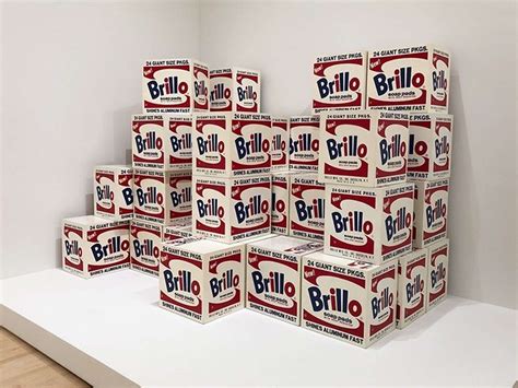 Andy Warhol's Brillo Box sculpture next to a real Brillo box in a gallery setting, illustrating the indistinguishability between art and everyday objects.