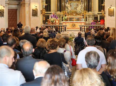 Foto della chiesa parrocchiale gremita durante la cerimonia funebre con fiori bianchi e partecipazione della comunità locale